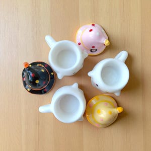 May include: A collection of whimsical, ceramic teacups and matching lids. The teacups are white with a handle, and the lids are decorated with polka dots and cone-shaped tops in black, pink, and yellow. The teacups are arranged on a light brown surface.