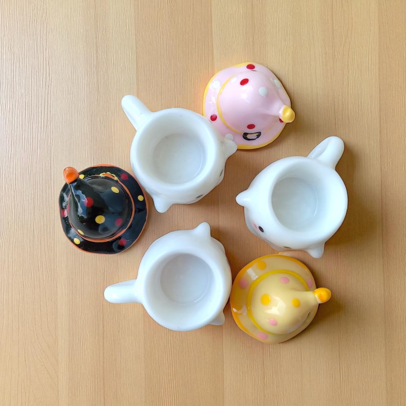 May include: A collection of whimsical, ceramic teacups and matching lids. The teacups are white with a handle, and the lids are decorated with polka dots and cone-shaped tops in black, pink, and yellow. The teacups are arranged on a light brown surface.