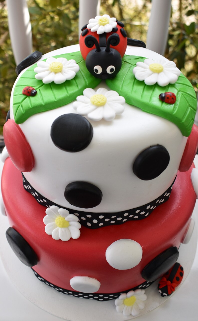 Pode incluir: Bolo de dois andares decorado com bolinhas vermelhas, brancas e pretas. O andar superior apresenta um desenho de folhas verdes com uma joaninha no topo. O andar inferior tem uma fita preta e branca.