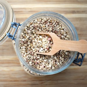 May include: A glass jar filled with small, beige and pink grains, with a wooden scoop inside. The jar has a metal lid and is sitting on a wooden surface.
