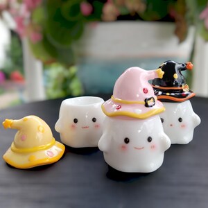 May include: A collection of white ceramic ghost figurines, each with a smiling face and rosy cheeks. Some ghosts wear colorful witch hats in yellow, pink, and black. The hats have star and dot accents. The figurines are on a dark surface.