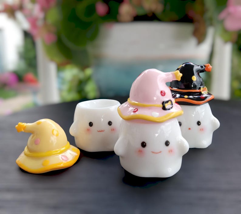 May include: A collection of white ceramic ghost figurines, each with a smiling face and rosy cheeks. Some ghosts wear colorful witch hats in yellow, pink, and black. The hats have star and dot accents. The figurines are on a dark surface.