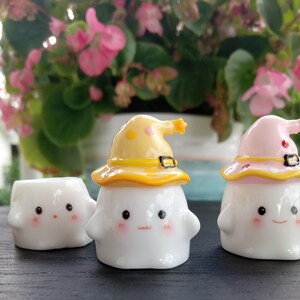 May include: Three white ceramic figurines with smiling faces and rosy cheeks. Two figurines wear yellow and pink witch hats with black buckles. The third figurine is hatless. The background features pink flowers and green leaves.