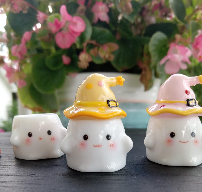 May include: Three white ceramic figurines with smiling faces and rosy cheeks. Two figurines wear yellow and pink witch hats with black buckles. The third figurine is hatless. The background features pink flowers and green leaves.