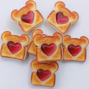 May include: Seven pieces of toast with a heart-shaped jelly center. The toast is golden brown with a light-colored crust. The jelly is a translucent red with small white specks. The toast pieces are arranged on a white surface.