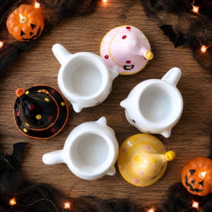 May include: Three white ceramic ghost-shaped mugs with handles, each accompanied by a removable conical lid. The lids feature polka dot patterns in pink, yellow, and black, designed to resemble witch hats sitting atop the ghost figures.