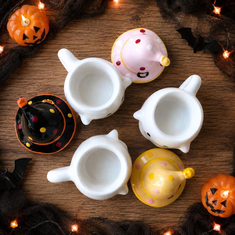 May include: Three white ceramic ghost-shaped mugs with handles, each accompanied by a removable conical lid. The lids feature polka dot patterns in pink, yellow, and black, designed to resemble witch hats sitting atop the ghost figures.