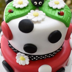Pode incluir: Um bolo de dois andares decorado com uma joaninha, margaridas brancas e bolinhas pretas e brancas. O andar inferior &eacute; vermelho e o andar superior &eacute; branco.
