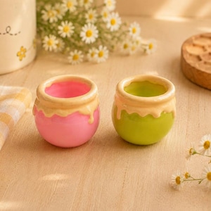 May include: Two small, round ceramic pots, one pink and one green, with a cream-colored glaze dripping from the rim. The pots are reminiscent of honey pots. Daisies and a wooden coaster are in the background.