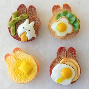 May include: Four whimsical, food-themed decorative items. Each piece is shaped like a slice of toast with a bunny ear design. The toppings include a fried egg, butter, lettuce, and a bunny with a carrot. The items are colorful and detailed.