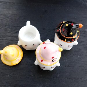 May include: A collection of ceramic ghost figurines, each with a unique witch hat design. One ghost is hatless, while others wear hats in yellow, pink, and black with polka dots. The figurines have smiling faces and are likely for Halloween.