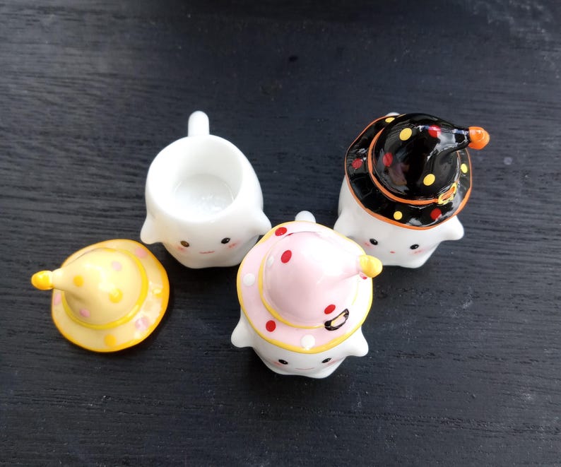 May include: A collection of ceramic ghost figurines, each with a unique witch hat design. One ghost is hatless, while others wear hats in yellow, pink, and black with polka dots. The figurines have smiling faces and are likely for Halloween.