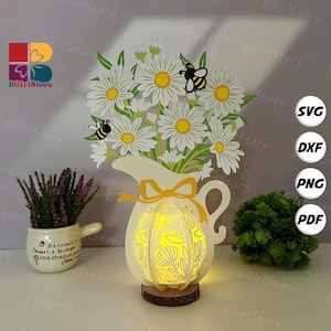 Könnte beinhalten: Dekorative Papierschnitt-Darstellung einer Vase mit Gänseblümchen und Bienen, von innen beleuchtet. Die Vase ist weiß mit einer gelben Schleife. Ein kleiner Topf mit Lavendel und eine grüne Pflanze sind auf beiden Seiten. Der Text "BGirlStore" ist enthalten.