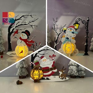 Peut inclure: Décorations de Noël en papier découpé représentant un bonhomme de neige, le Père Noël et un ours polaire avec un pingouin. Chaque scène comprend une lanterne lumineuse. Les scènes incluent des paysages enneigés et des arbres d'hiver.