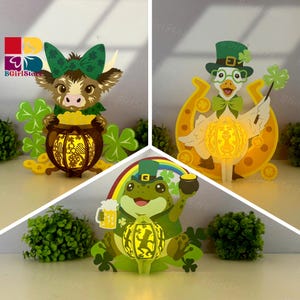 Op de afbeelding: Drie papieren uitgesneden decoraties met het thema St. Patrick's Day. Eén toont een koe met een pot goud, een ander een gans met een klavertje vier en de laatste een kikker met bier en een pot goud. Elk heeft een gloeiende lantaarn.