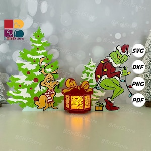 Może przedstawiać: Świąteczna dekoracja wycinana laserowo z Grinchem, Maksem i świecącym pudełkiem na prezent. Grinch ma na sobie strój Świętego Mikołaja i trzyma światełka. Obecne są również choinka i renifer.