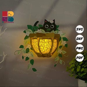 May include: A hanging paper lantern with a cat design. The lantern is yellow with a floral pattern and emits a warm glow. A black cat with a flower on its head sits above the lantern, surrounded by green leaves and white flowers. The image includes the text "SVG", "DXF", "PNG", and "PDF".
