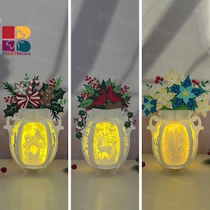 May include: Three illuminated paper lanterns with intricate cut-out designs. Each lantern features a different festive arrangement: a Christmas theme with a gingerbread man, cardinals, and poinsettias. The lanterns are white with a warm yellow glow.