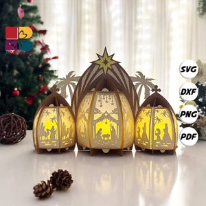 May include: A brown and white paper Christmas nativity scene with three illuminated lanterns. The lanterns feature cut-out silhouettes of the nativity scene. The scene includes a star, palm trees, and the text "SVG, DXF, PNG, PDF".