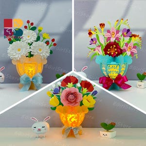 Könnte beinhalten: Drei Papierblumenarrangements in Vasen. Eines zeigt weiße Gänseblümchen, ein anderes eine Mischung aus roten und lila Blumen und das dritte eine rosa Rose. Jede Vase hat eine Schleife und wird von innen beleuchtet. Der Text "Best Mom Ever" ist sichtbar.
