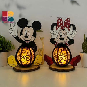 May include: Two decorative Mickey and Minnie Mouse lanterns. The black and yellow figures are on wooden bases, with cut-out designs that glow with warm light. Mickey is giving a peace sign. Minnie has a red bow.
