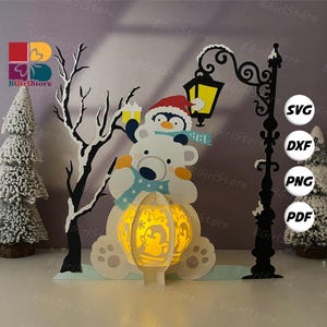 Puede incluir: Una escena de manualidades con temática invernal con un oso polar y un pingüino. El oso sostiene una linterna brillante con un diseño de pingüino. También hay una farola negra y un árbol. La escena está ambientada sobre un fondo morado claro. La imagen incluye iconos de tipo de archivo SVG, DXF, PNG y PDF.