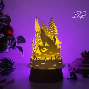 May include: A white paper lamp with a wolf howling at the moon in a forest scene. The lamp is lit from within, casting a warm glow. The lamp is on a wooden base.