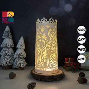 Peut inclure: Une lanterne en papier cylindrique avec une scène de la Nativité découpée, comprenant Marie, Joseph et l'enfant Jésus, est posée sur une base en bois. La lanterne a un dessus décoratif en forme de couronne. L'image comprend également de petits arbres de Noël et le logo BGirlStore.