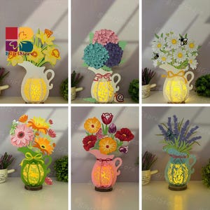 May include: A selection of decorative paper flower arrangements in the shape of a jug. Each jug is illuminated from within, displaying intricate cut-out designs. The flowers are various colours, including yellow, pink, and purple.