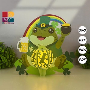 Puede incluir: Una manualidad de papel con temática del Día de San Patricio que presenta una rana verde con un sombrero de duende, sosteniendo una jarra de cerveza y una olla de oro. Un arcoíris se arquea detrás de la rana, y una linterna brillante está al frente. Incluye el texto "SVG, DXF, PNG, PDF".