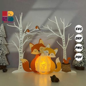 May include: A layered paper art scene featuring two orange foxes embracing, with a glowing lantern between them. White birch trees, birds, and a squirrel on a stump complete the woodland design. The image includes the text "SVG, DXF, PNG, PDF".