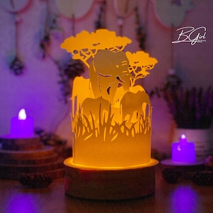 May include: A yellow paper lamp with a silhouette of elephants and trees. The lamp is lit and casting a warm glow. The lamp is on a wooden base.