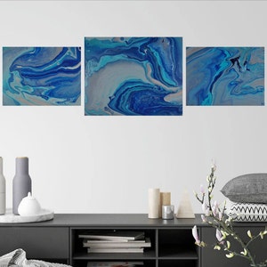 May include: Three abstract paintings in shades of blue and white with a marbled effect. The paintings are hung on a white wall above a dark gray console table.