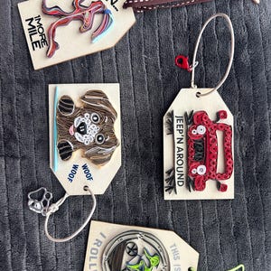 May include: Wooden tags with paper quilling art. One tag says "RUN... 1 MORE MILE" with a colorful abstract design. Another features a dog with "WOOF WOOF". A red Jeep is on a third tag with "JEEP 'N AROUND". The last tag says "THIS IS HOW... /ROLL!!!" with a bicycle.