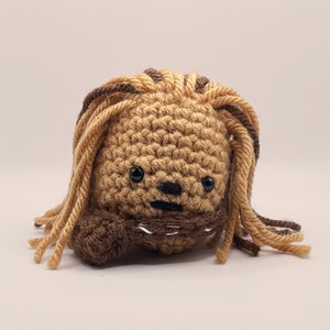 May include: A crocheted plush toy resembling Chewbacca from Star Wars. The toy is brown and tan with a black nose and eyes. It has long brown and tan yarn hair.