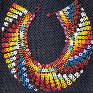 May include: A vibrant beaded necklace in a U-shape, showcasing a spectrum of colours: red, yellow, green, blue, and white. The beads are arranged in a geometric pattern, creating a striking visual effect. The necklace is likely handmade, a unique piece of jewellery.