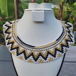 May include: A beaded necklace with a geometric pattern in black, white, and gold. The necklace is displayed on a white mannequin bust. The design features a zig-zag pattern, creating a bold and eye-catching accessory.