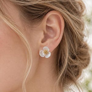 以下が含まれることがあります： 3枚の白い半透明の花びらと、質感のある金色の中心部を持つ小さな花形のスタッドピアス。ピアスはポスト式の留め具で耳たぶに固定されており、繊細でシンプルな花の形をしたデザインが特徴です。