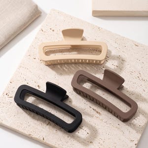 Puede incluir: Tres pinzas rectangulares para el cabello con acabado mate. Una pinza es de color beige, otra marrón y otra negra. Cada pinza presenta un diseño de marco abierto ancho con dientes entrelazados en el borde inferior y un mecanismo de resorte.