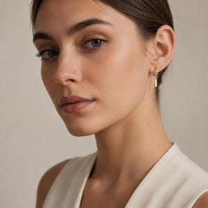 May include: A silver hoop earring featuring a small, vertical rectangular bar charm suspended from the bottom. The earring is worn in the earlobe, showcasing a polished metallic finish that reflects light, creating a clean and simple aesthetic.
