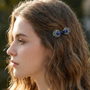 May include: A decorative hair clip featuring a bow design with a dark silver finish. The bow is adorned with two oval-shaped, deep blue gemstones. The clip is set against a backdrop of wavy, light brown hair.