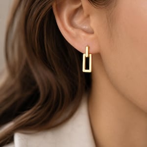 May include: A gold-toned earring featuring a small rectangular post connected to a larger, vertically oriented rectangular drop. The piece has a polished, smooth metallic finish and a simple, geometric design that hangs directly from the earlobe.