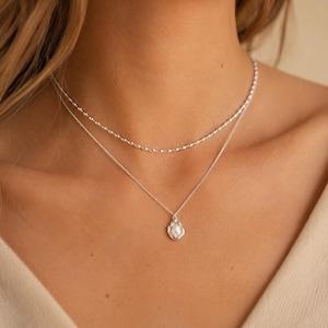 May include: A layered silver necklace set consisting of two separate chains. The top chain features a delicate beaded texture, while the bottom chain holds a small, irregular silver pendant centered with a single white pearl-like accent.