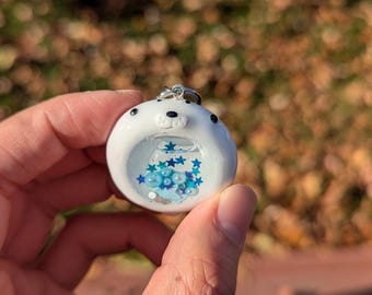 May include: A white, round pendant with a seal face design. The pendant has a clear center filled with blue stars and beads. A silver-colored metal loop is attached at the top.