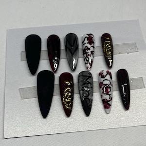 Pode incluir: Um conjunto de dez unhas postiças longas em forma de amêndoa. As unhas são pintadas com vários desenhos, incluindo preto liso, bordô, cinza com chevron e branco com detalhes florais vermelhos. Algumas unhas apresentam detalhes dourados e as palavras "Mine" e "Sorry".