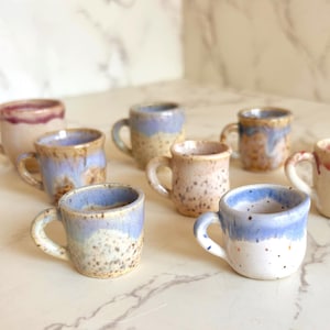 May include: A collection of eight miniature ceramic mugs featuring various speckled glazes in shades of blue, cream, and reddish-brown. Each small cup has a rounded body, a single handle, and a unique hand-finished appearance with distinct color variations.