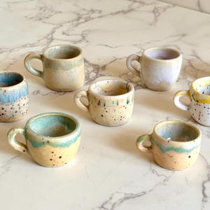 May include: A collection of eight small ceramic cups with handles, each featuring a unique glaze finish. The cups display various colors including blue, yellow, tan, and white, with some showing speckled textures, drip patterns, and smooth gradients.