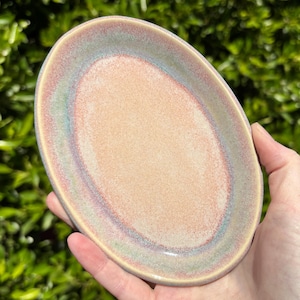 May include: An oval-shaped ceramic dish with a peach-colored center and a rim with iridescent hues of blue, green, and purple. The dish is held in a hand against a backdrop of green foliage. Ideal for serving small portions.