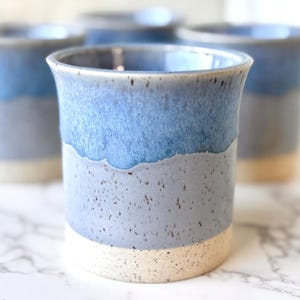 May include: Four handmade ceramic tumblers with a layered blue and gray glaze. The top section is a vibrant blue, transitioning to a speckled gray, and ending with a natural, unglazed base. These drinking cups are perfect for water, juice, or other beverages.