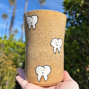 May include: A tan ceramic tumbler with a speckled finish, adorned with smiling white tooth illustrations. The cylindrical cup is held in a hand, with a backdrop of green foliage and a blue sky.